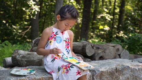 A girl sits on a rock wall with a paint palette by her side, painting on a paper on her lap