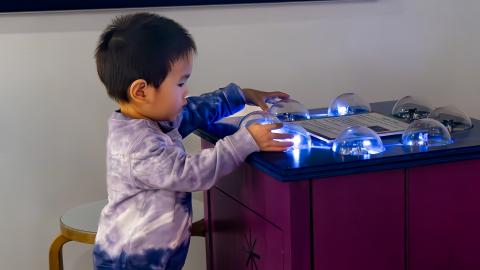 a young boy stands in front of a tabletop exhibit with a circle of blinking lights