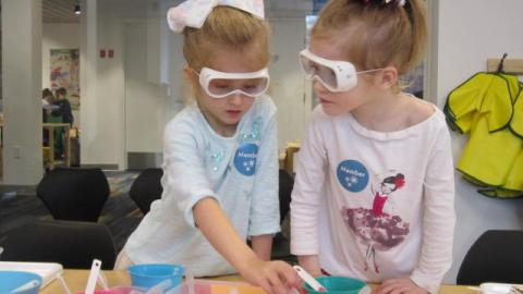 Two girls wearing safety goggles stand at a table with several small colorful bowls