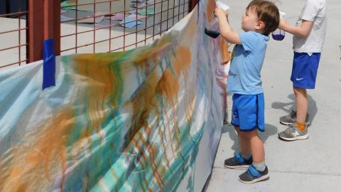 A white sheet full of paint splatter is taped to a fence where two boys are spraying it with paint from spray bottles.