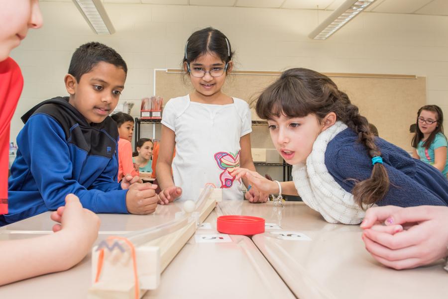 A small group of children watches a marble roll down a wooden track