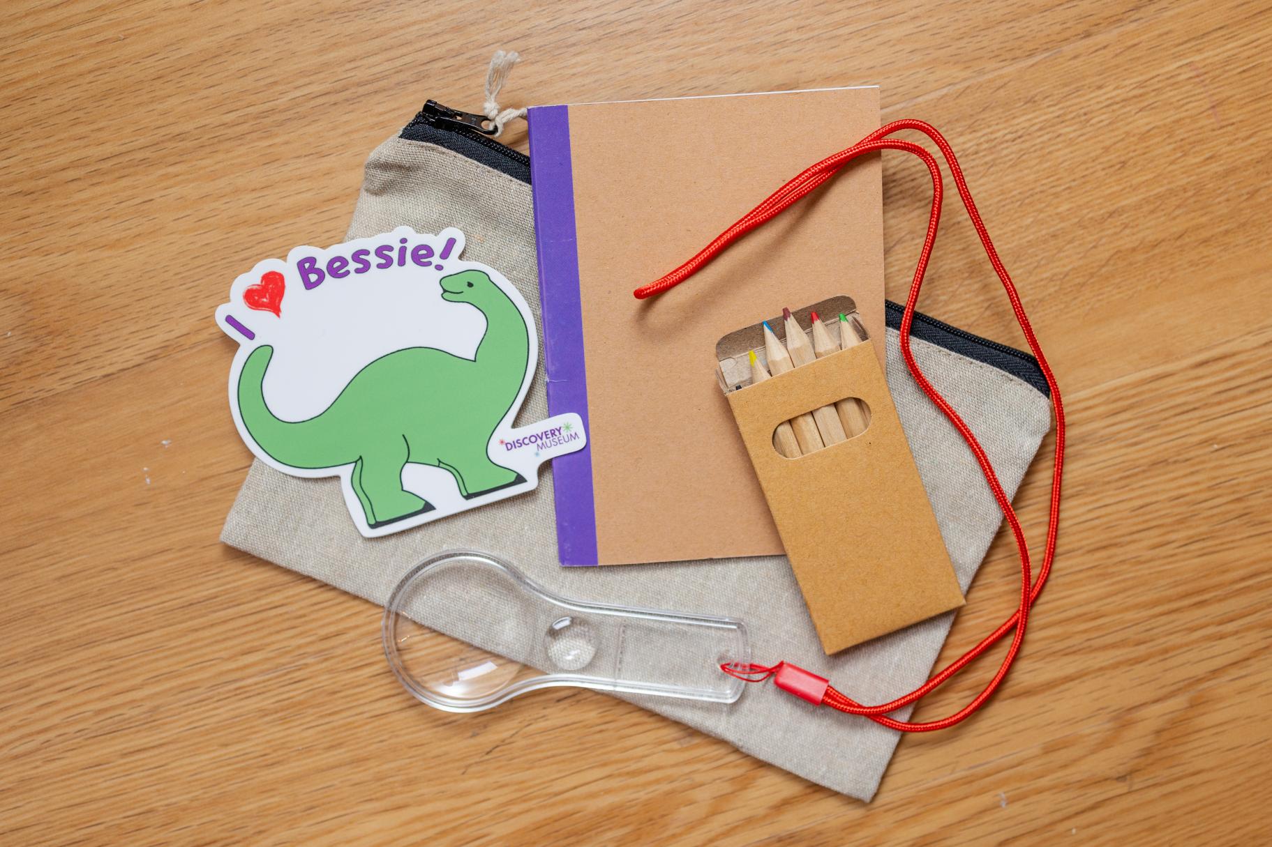 a small notebook, magnifying glass, pencil and dino sticker on top of a small canvas bag